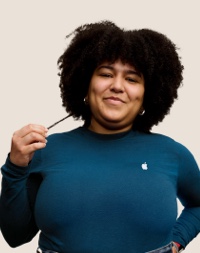 Portrait d’une personne de l’équipe de vente au détail d’Apple souriant; elle a les cheveux foncés, longs et bouclés.