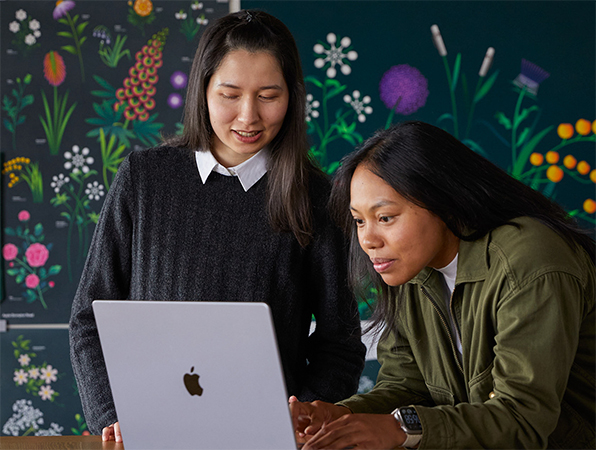 سيدتان من أعضاء فريق التسويق في Apple تعملان معاً على جهاز MacBook أمام ملصقات من تصميم فريقهما.