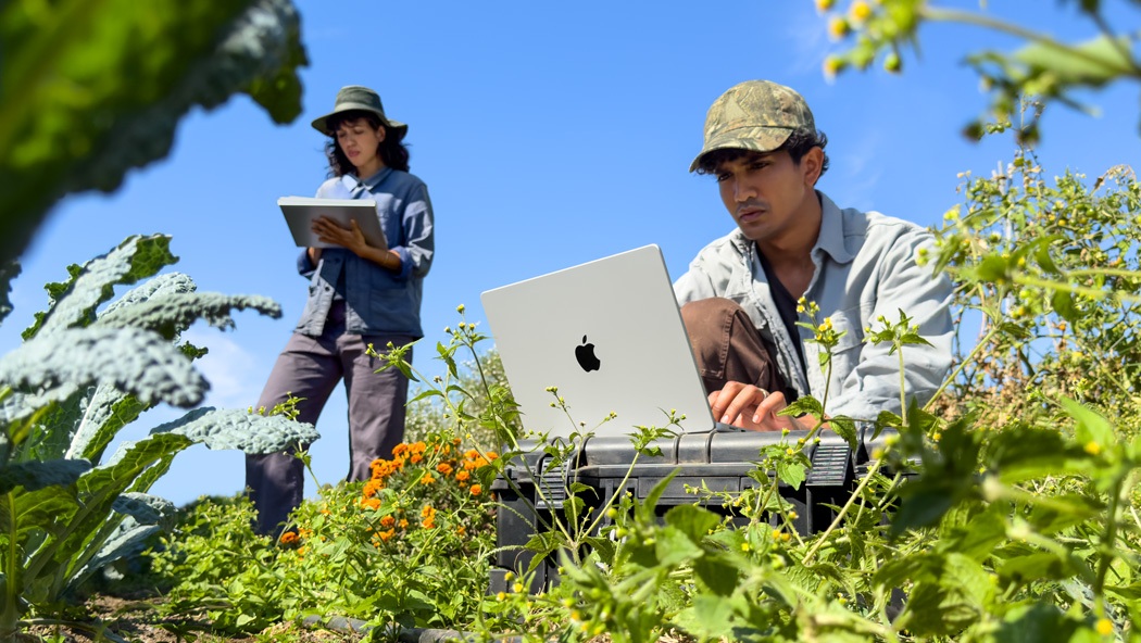 Two people working outdoors in a garden, one using an unplugged silver MacBook Pro and the other holding a notepad